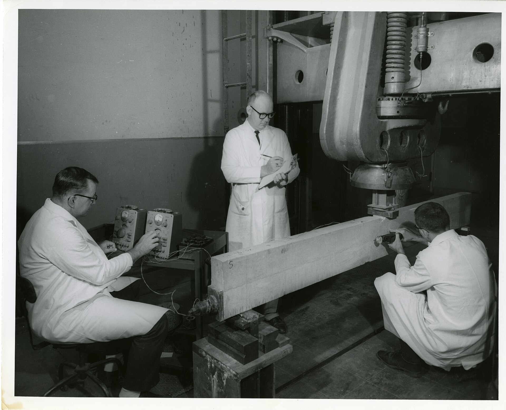 Three researchers examine a concrete beam in a structural testing machine.
