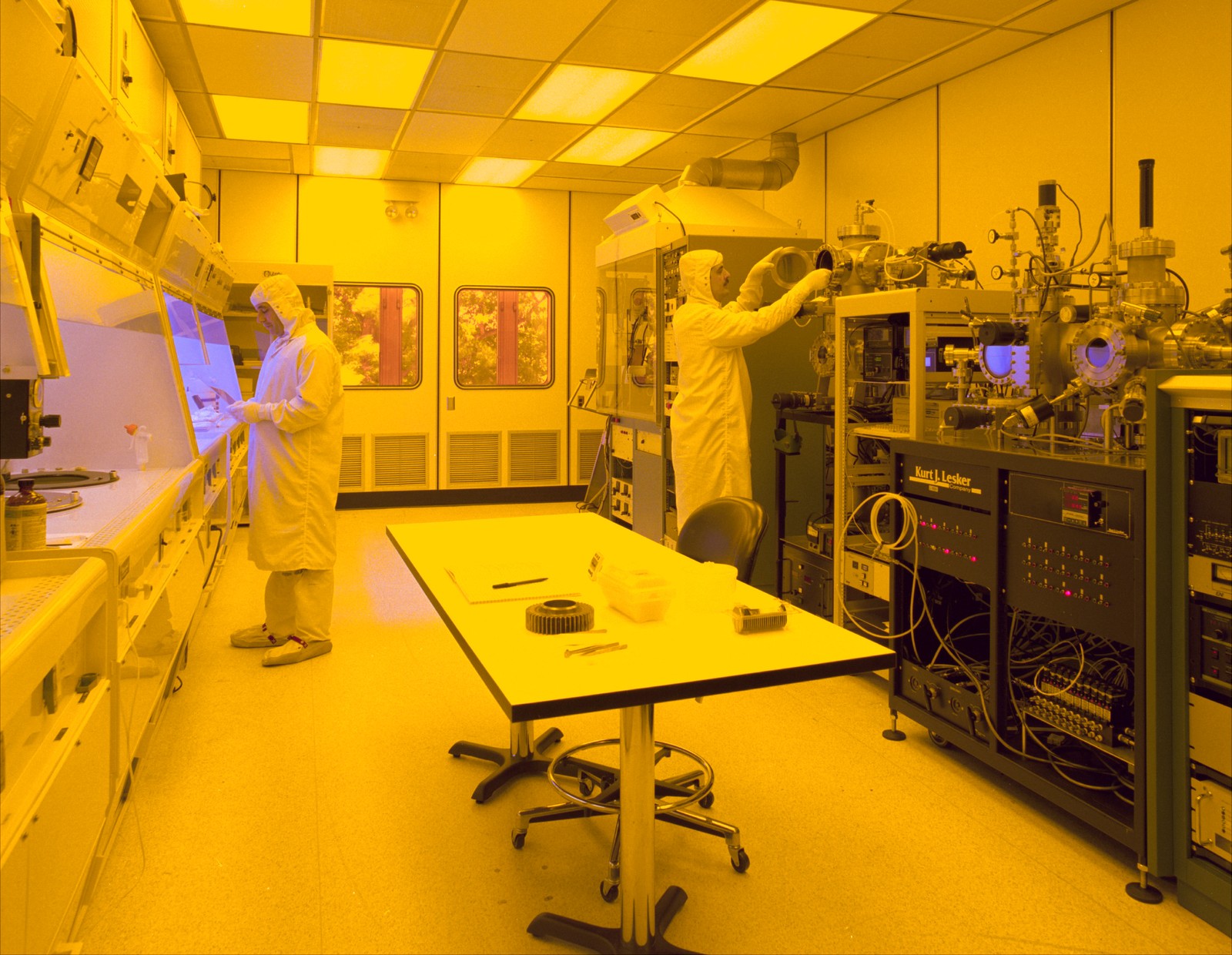 Two people in white hooded suits work at equipment in a yellow-lit cleanroom, with a table in the foreground.