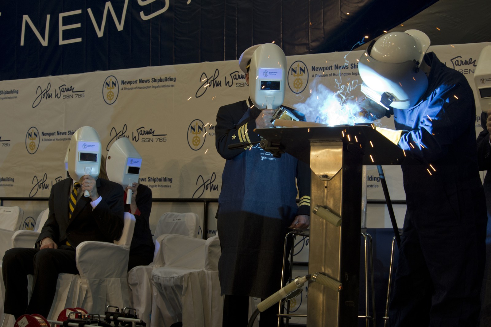 Sparks fly as a welder bends over a steel plate while ceremony attendees watch through protective welding shields.
