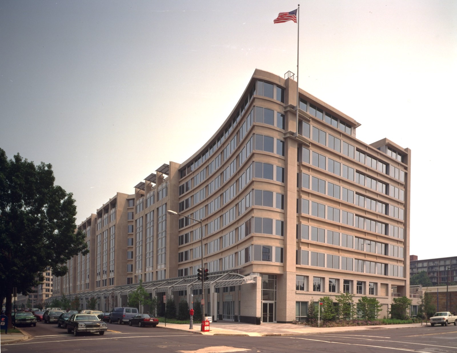A tan office building with a curved facade and rows of windows stands at a street intersection, with a U.S. flag overhead.