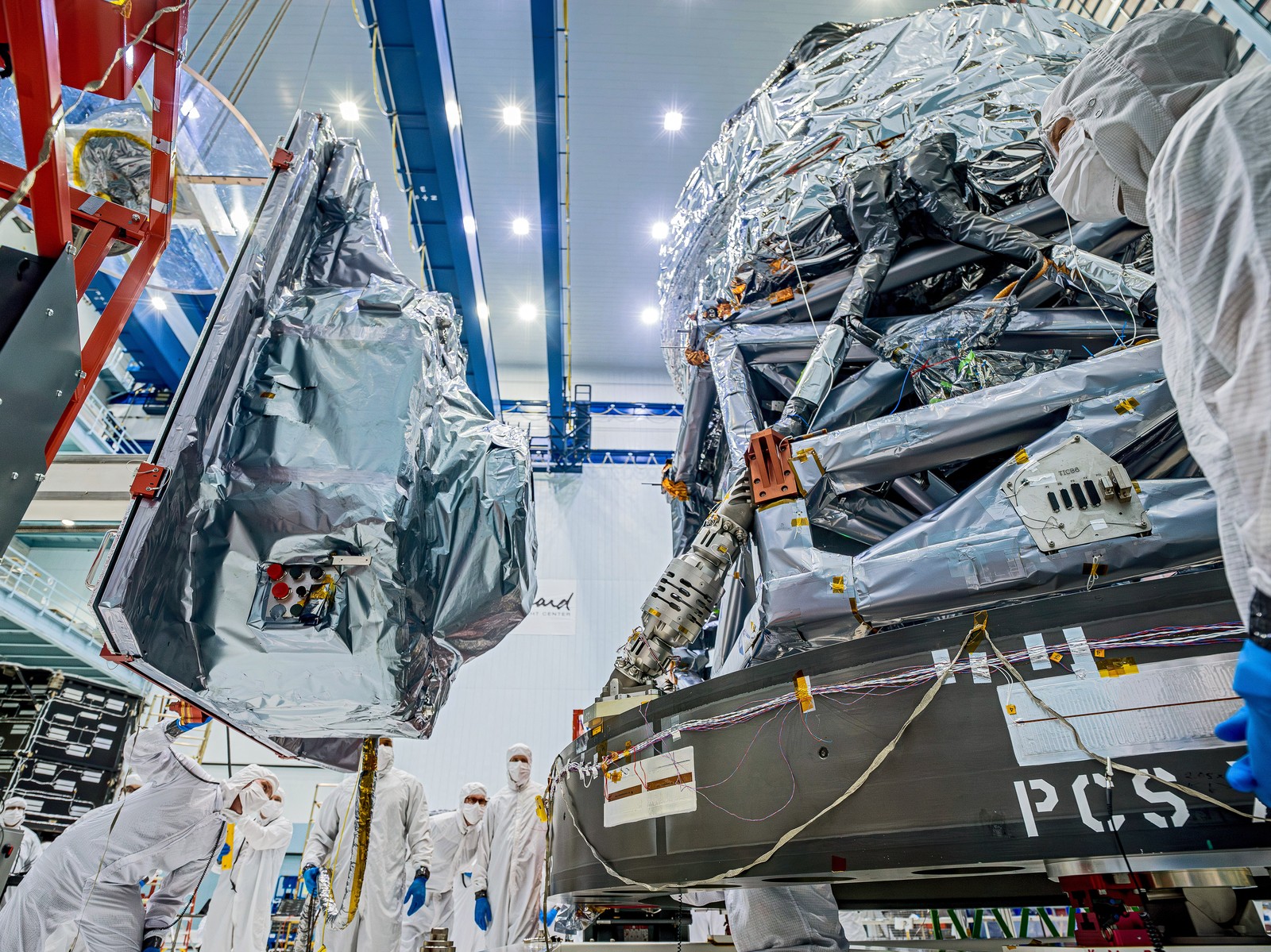 Technicians in white clean-room suits work beside foil-wrapped Roman telescope hardware, with the Wide Field Instrument suspended at left.