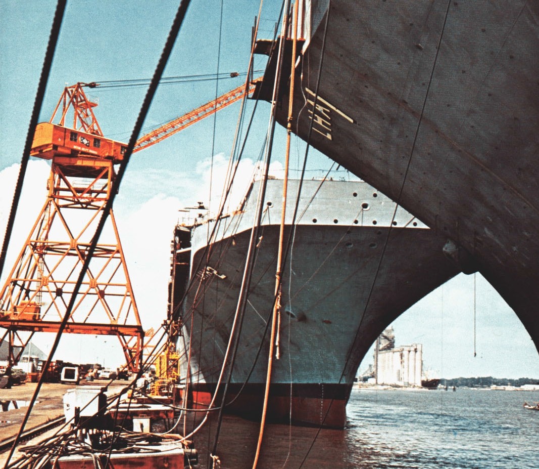 Two large ship hulls beside a dock, with an orange crane at left and cables crossing the foreground.